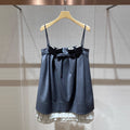A dark blue-gray plaid mini dress with thin shoulder straps, a large black bow at the waist, and a white pleated hem, hanging on a wooden hanger against a textured beige wall.