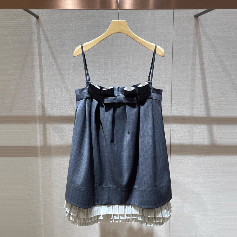 A dark blue-gray plaid mini dress with thin shoulder straps, a large black bow at the waist, and a white pleated hem, hanging on a wooden hanger against a textured beige wall.