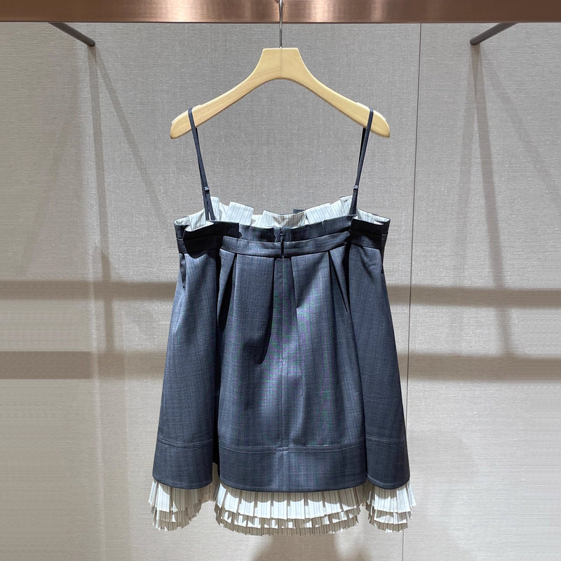 A dark blue-gray mini skirt with thin shoulder straps, a ruffled waistband, pleated front, and a white ruffled hem, hanging on a wooden hanger against a neutral wall.