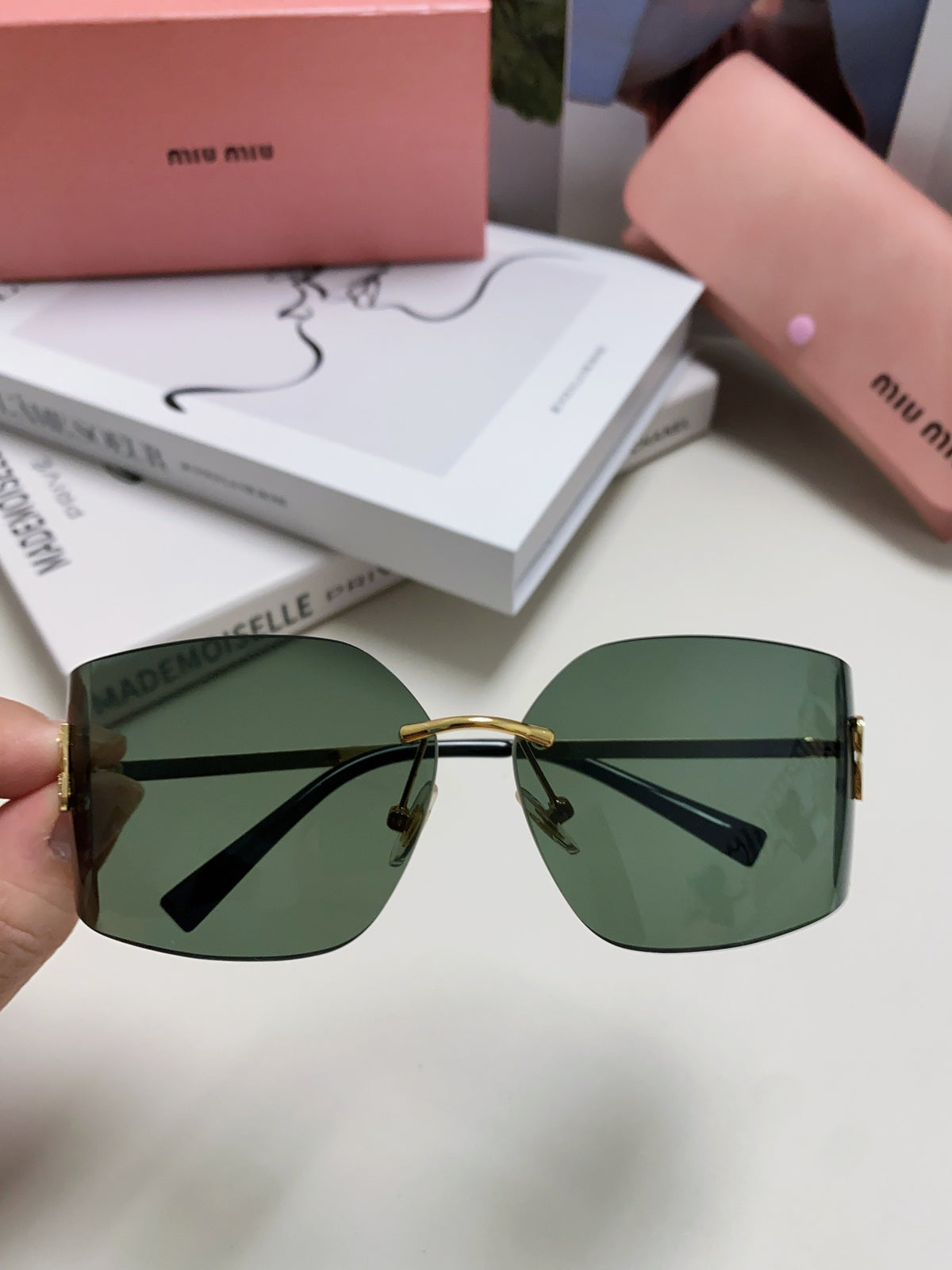 Miu miu sunglasses displayed with a pink case and books in the background, showcasing stylish green lenses.