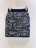 A short knit pencil skirt with a dark navy blue base and a gray and white abstract floral or medallion pattern, hanging from a clear plastic hanger with two clips against a plain off-white wall.