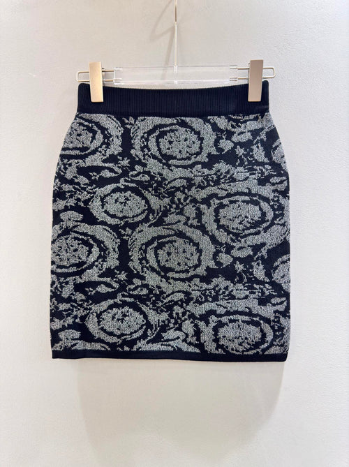A short knit pencil skirt with a dark navy blue base and a gray and white abstract floral or medallion pattern, hanging from a clear plastic hanger with two clips against a plain off-white wall.